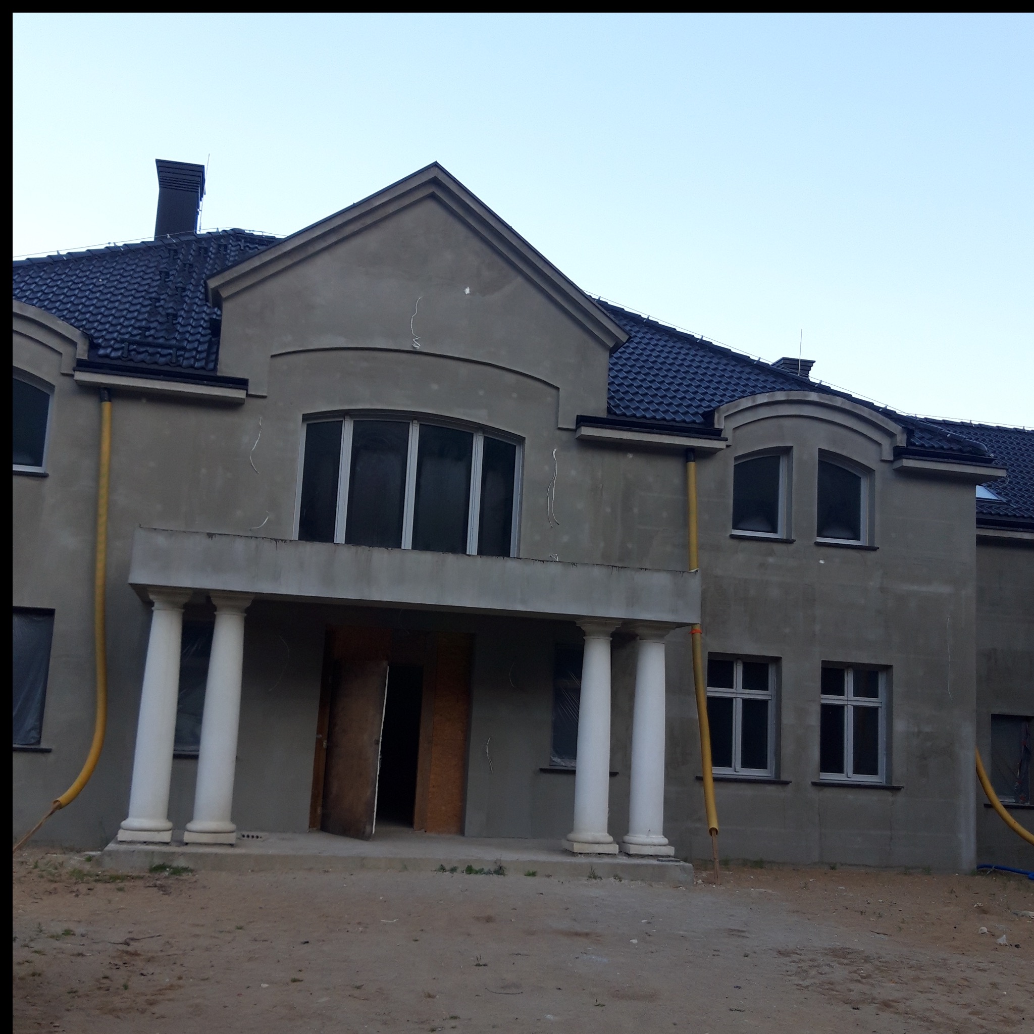 Surowy dom z kolumnami przed wejściem, w trakcie prac wykończeniowych. Widoczne rury spustowe i piaskowe podłoże.