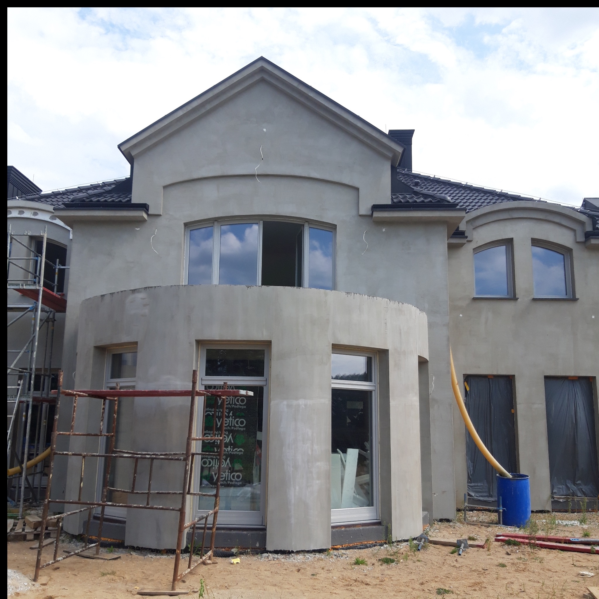 Dom w trakcie prac wykończeniowych elewacji, z widocznymi rusztowaniami i materiałami budowlanymi. Ściany pokryte szarą warstwą, okna częściowo zabezpieczone folią.