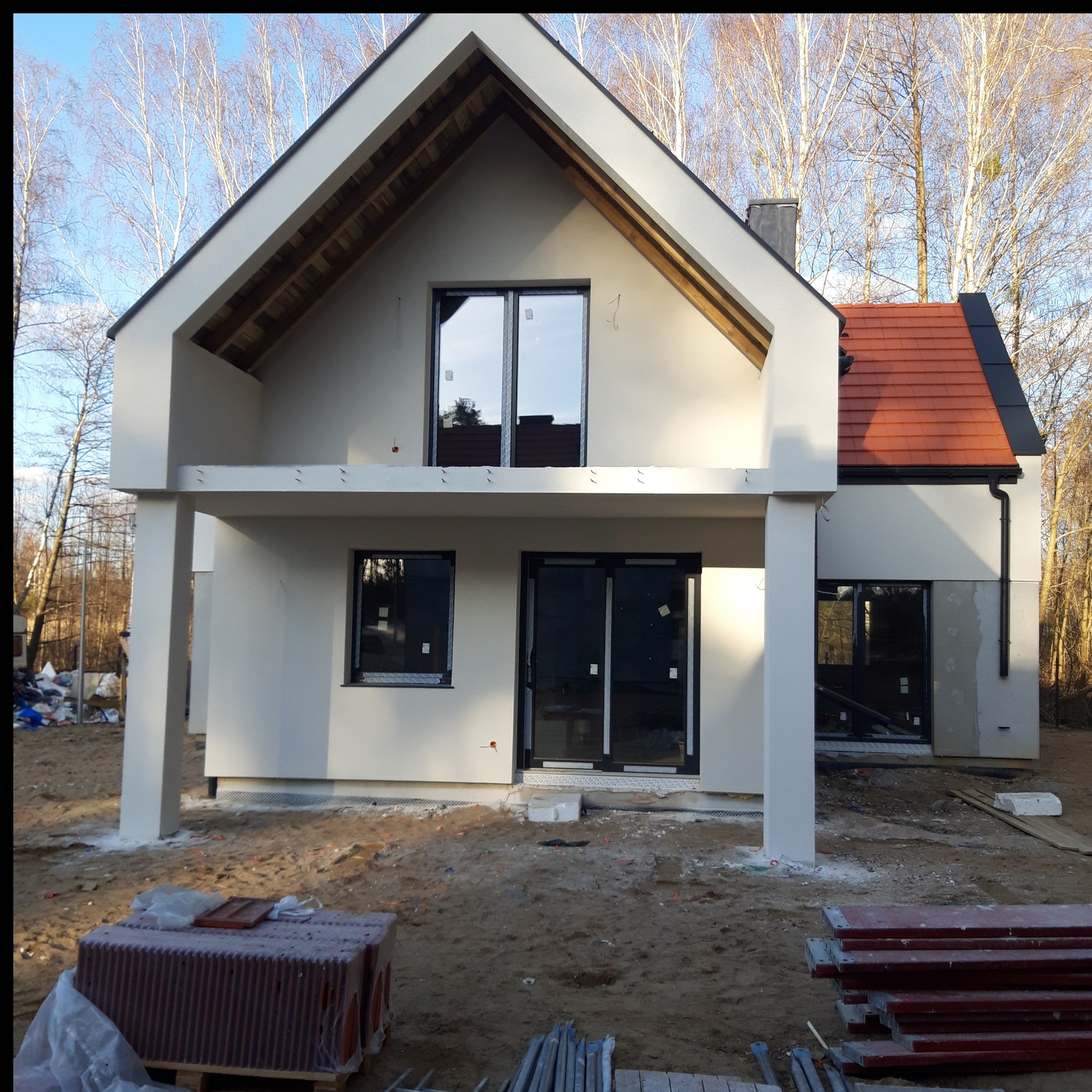 Nowoczesny dom jednorodzinny w trakcie wykończenia elewacji zewnętrznej. Widoczne okna z czarnymi ramami, czerwona dachówka i elementy konstrukcyjne balkonu.