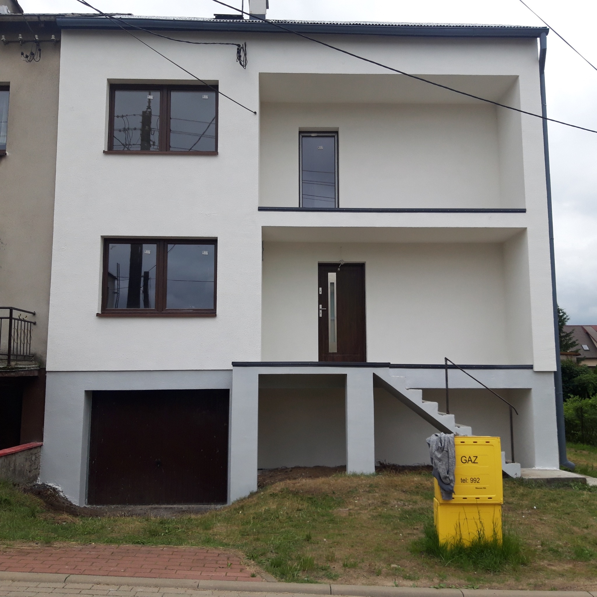 Dwukondygnacyjny dom po renowacji z białymi ścianami, ciemnobrązowymi ramami okiennymi i drzwiami. Widoczna skrzynka gazowa i schody wejściowe.