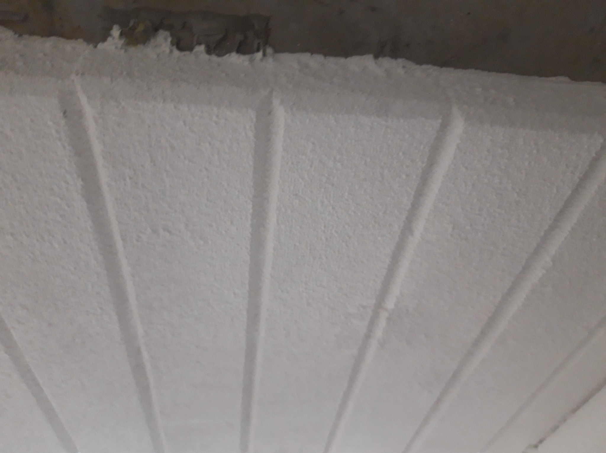 Biała, świeżo położona gładź na suficie z widocznymi, równoległymi liniami. Tekstura gładzi jest delikatnie chropowata, a linie tworzą subtelny wzór.