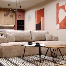 Agata Kębłowska Strefa Projektowania - Jasny salon z beżową sofą, stolikami kawowymi i dywanem w paski. W tle zabudowa kuchenna w odcieniach brązu i różu, oraz abstrakcyjne obrazy na ścianie.