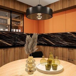 Agata Kębłowska Strefa Projektowania - Nowoczesna kuchnia z drewnianymi frontami, czarnym blatem imitującym marmur i pomarańczowymi akcentami. Okrągły stół z dekoracjami na pierwszym planie.