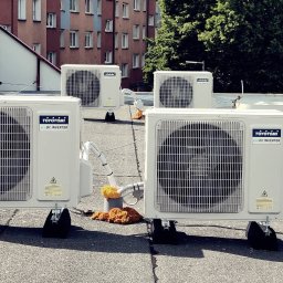 KlimAlex - Pompy ciepła, Klimatyzacja, Ogrzewanie podłogowe - Cztery jednostki zewnętrzne klimatyzacji Toyotomi Eco DC Inverter na dachu budynku z cegły, widoczne elementy instalacji. Tło: budynek mieszkalny i zieleń.