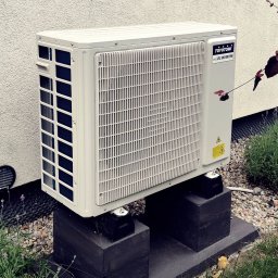 KlimAlex - Pompy ciepła, Klimatyzacja, Ogrzewanie podłogowe - Biała jednostka zewnętrzna klimatyzacji Toyotomi eco DC Inverter, zamontowana na betonowych podstawach, na tle jasnej ściany i zieleni.