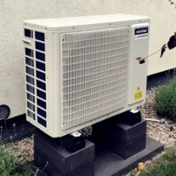 Biała jednostka zewnętrzna klimatyzacji Toyotomi eco DC Inverter, zamontowana na betonowych podstawach, na tle jasnej ściany i zieleni.