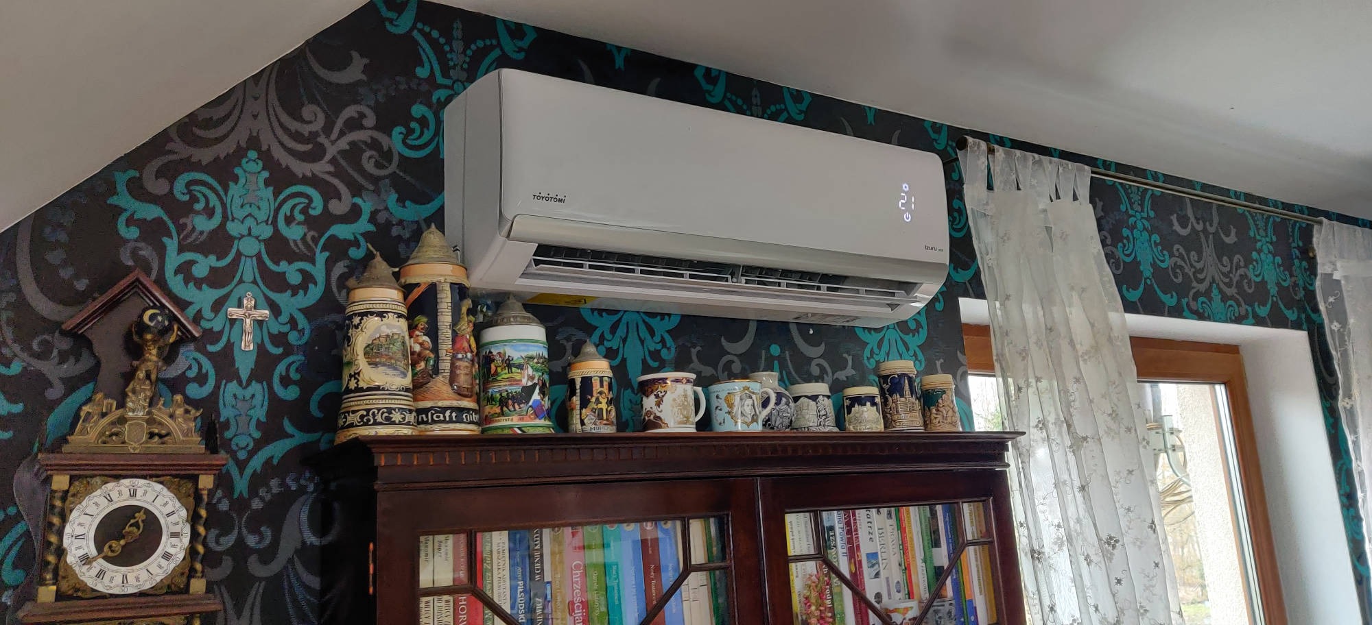 Biały klimatyzator Toyotomi zamontowany na ścianie z wzorzystą tapetą, nad drewnianym regałem z książkami i kolekcją ceramicznych kufli.