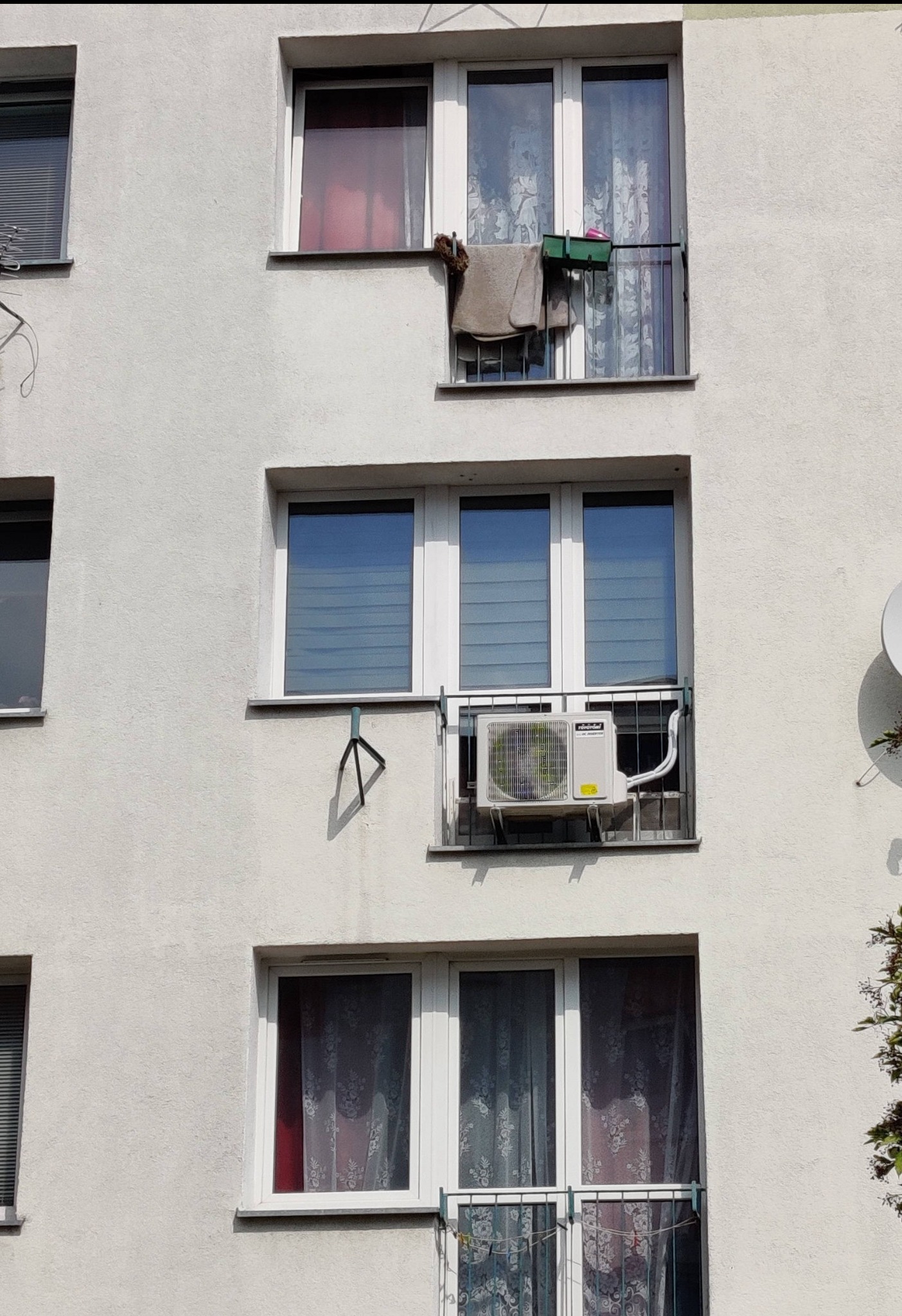 Biała ściana bloku z trzema oknami; na środkowym oknie zamontowany klimatyzator na balkonie z metalową barierką, widoczne rury odprowadzające.