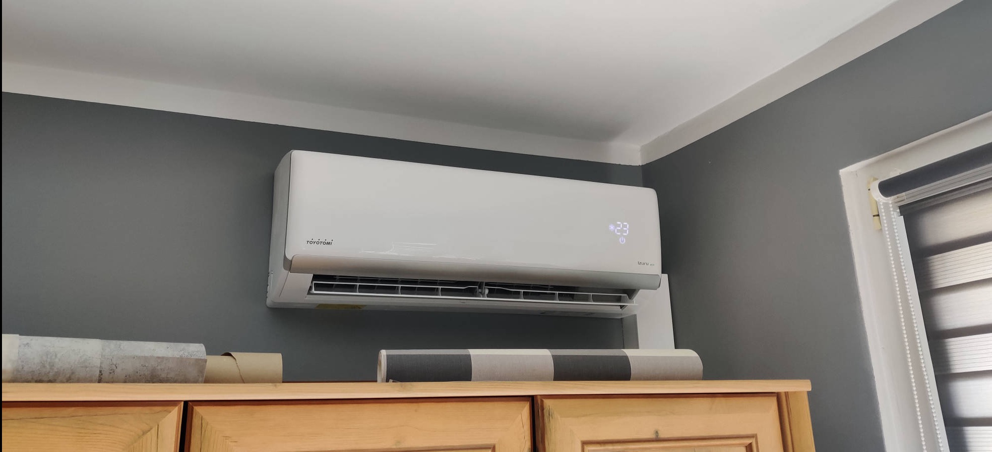 Biały klimatyzator Toyotomi na szarej ścianie, z wyświetlaną temperaturą 23 stopnie Celsjusza, zamontowany nad drewnianą komodą w pomieszczeniu z roletami.