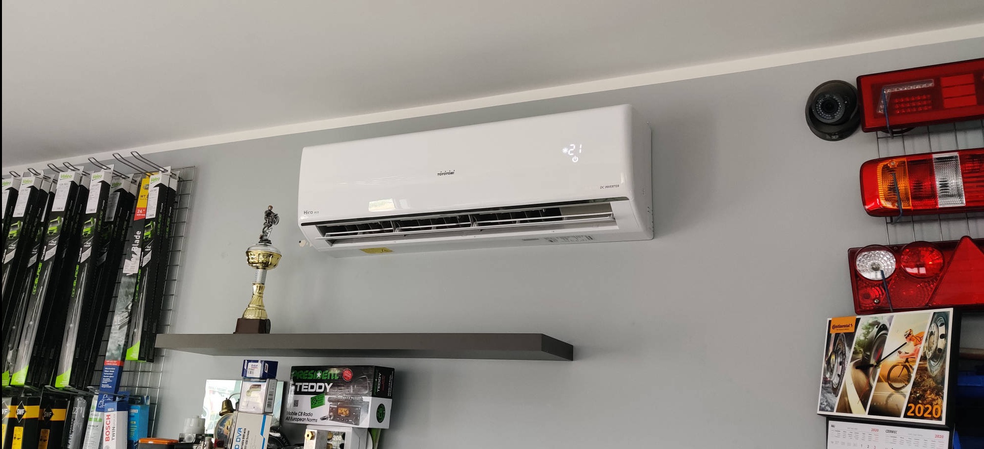 Biały klimatyzator Toyotomi na szarej ścianie w sklepie, pod nim półka z trofeum i pudełkami, obok akcesoria samochodowe i kamery. Widoczny wyświetlacz z temperaturą.