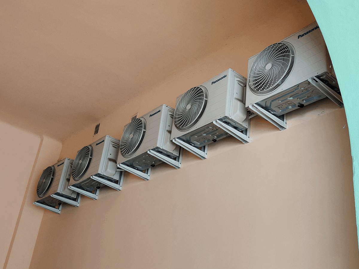 Rząd klimatyzatorów Panasonic zamontowanych na ścianie w jasnym pomieszczeniu. Widoczne wsporniki i detale instalacji. Ujęcie z dołu, podkreślające regularność rozmieszczenia.