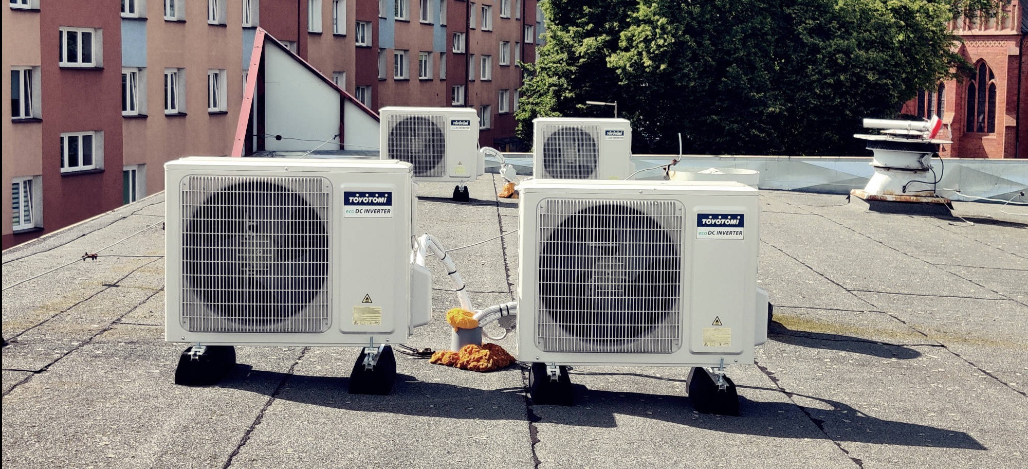 Cztery jednostki zewnętrzne klimatyzacji Toyotomi Eco DC Inverter na dachu budynku z cegły, widoczne elementy instalacji. Tło: budynek mieszkalny i zieleń.