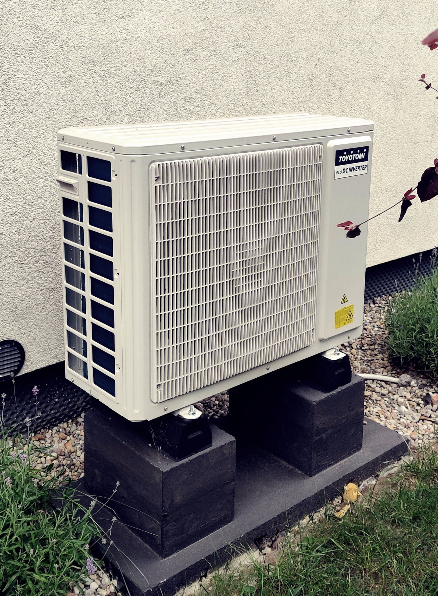 Biała jednostka zewnętrzna klimatyzacji Toyotomi eco DC Inverter, zamontowana na betonowych podstawach, na tle jasnej ściany i zieleni.