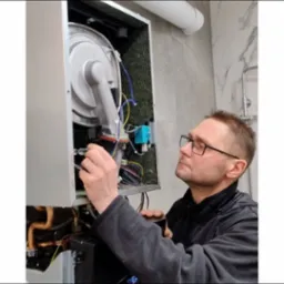 Mężczyzna w okularach serwisuje piec gazowy w łazience z marmurowymi płytkami. Widoczne przewody i elementy pieca.