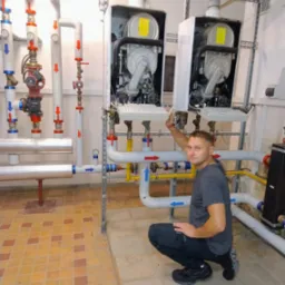 Instalator w szarym T-shircie kuca przed dwoma piecami gazowymi i instalacją z rur oznaczonych strzałkami w pomieszczeniu z kafelkowaną podłogą.