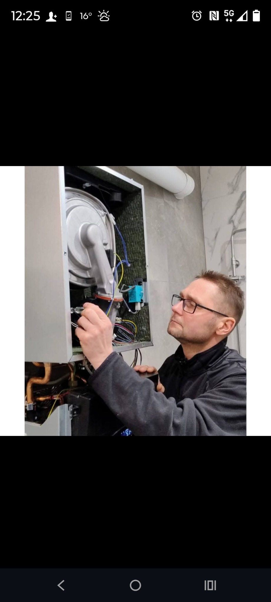 Mężczyzna w okularach serwisuje piec gazowy w łazience z marmurowymi płytkami. Widoczne przewody i elementy pieca.