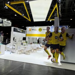 M&s bud construction spółka zoo - Stoisko targowe Solax Power z grafiką piłkarzy BVB Dortmund, białe meble i żółte akcenty. Widoczne logo Nord Technology. Oświetlenie sufitowe.