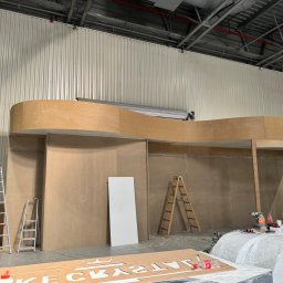 M&s bud construction spółka zoo - Konstrukcja z płyt drewnopochodnych w hali, widoczne rusztowania i narzędzia. Etap budowy stoiska targowego lub scenografii.