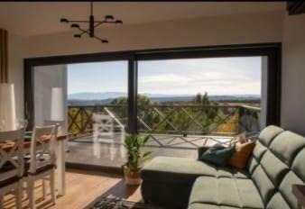 Wnętrze salonu z dużym oknem balkonowym i widokiem na krajobraz. Nowoczesny design, drewniana podłoga, zielona sofa i dekoracje. W tle góry i błękitne niebo.