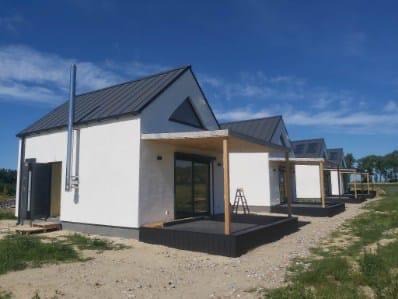 Rząd nowoczesnych, białych domków z szarymi dachami i drewnianymi tarasami w słoneczny dzień. Widoczna drabina oparta o jeden z domków i komin.