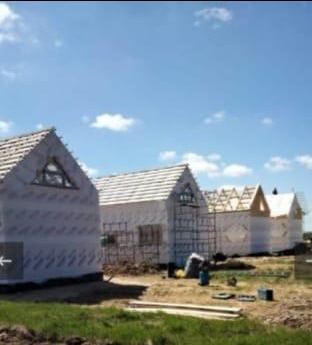 Wznoszenie szeregu domów w stanie surowym otwartym, z widoczną białą membraną izolacyjną i drewnianą konstrukcją dachu, na tle błękitnego nieba.