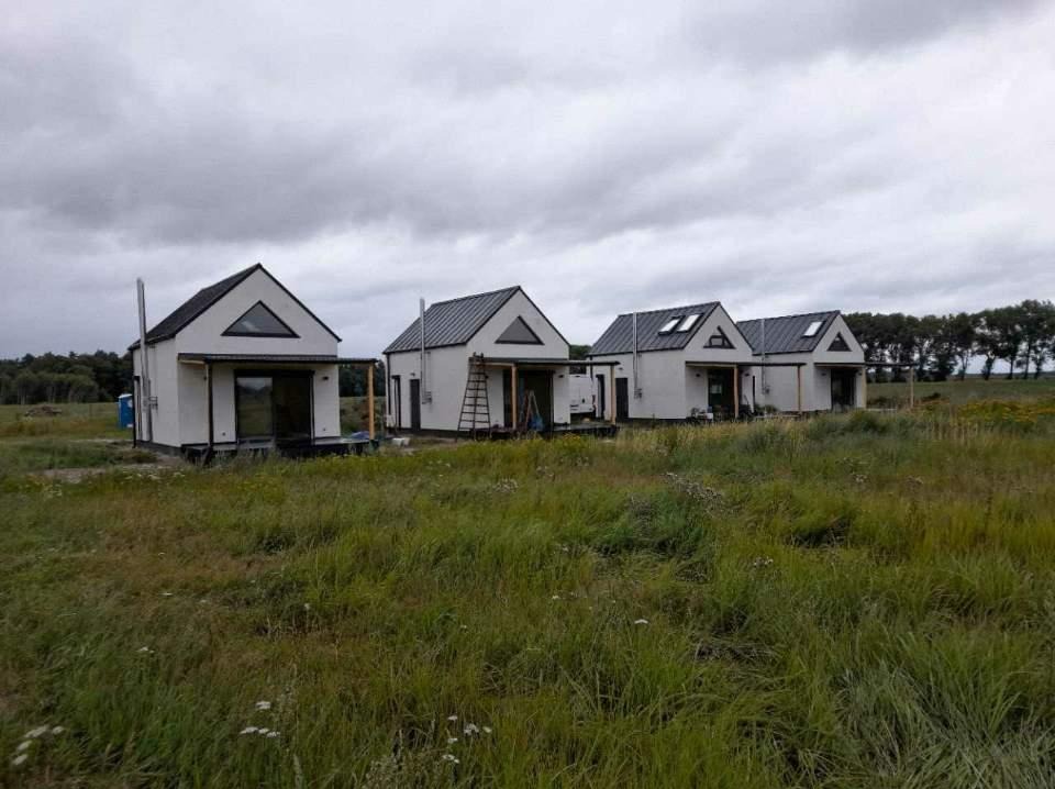 Cztery identyczne, nowoczesne, białe domki z czarnymi dachami, ustawione w rzędzie na łące pod pochmurnym niebem. Widoczna drabina oparta o jeden z domków.