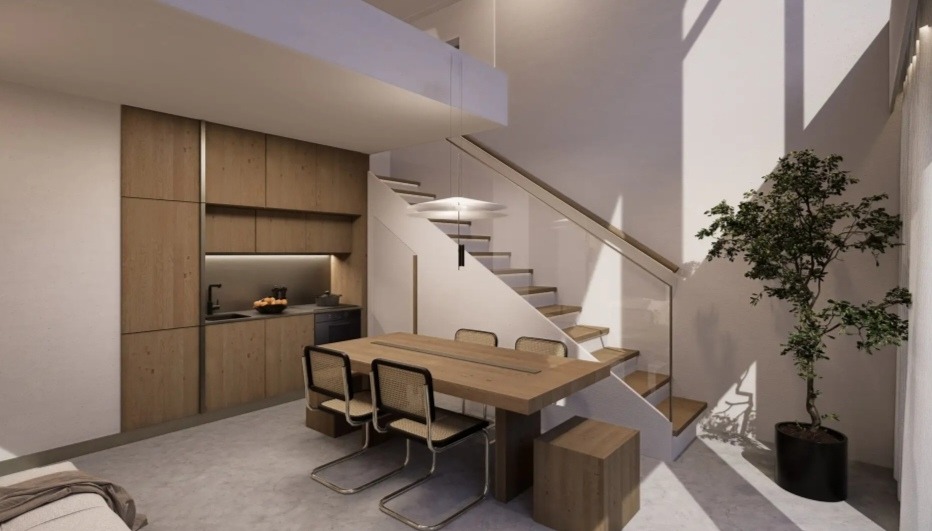 Jasne, nowoczesne wnętrze: drewniana zabudowa kuchenna, stół z krzesłami, schody z balustradą i roślina doniczkowa, oświetlone wiszącą lampą.