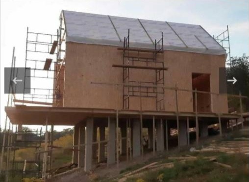 Dom w trakcie budowy, ściany obłożone płytami, widoczne rusztowania i podpory, krajobraz pagórkowaty w tle, etap surowy zamknięty.