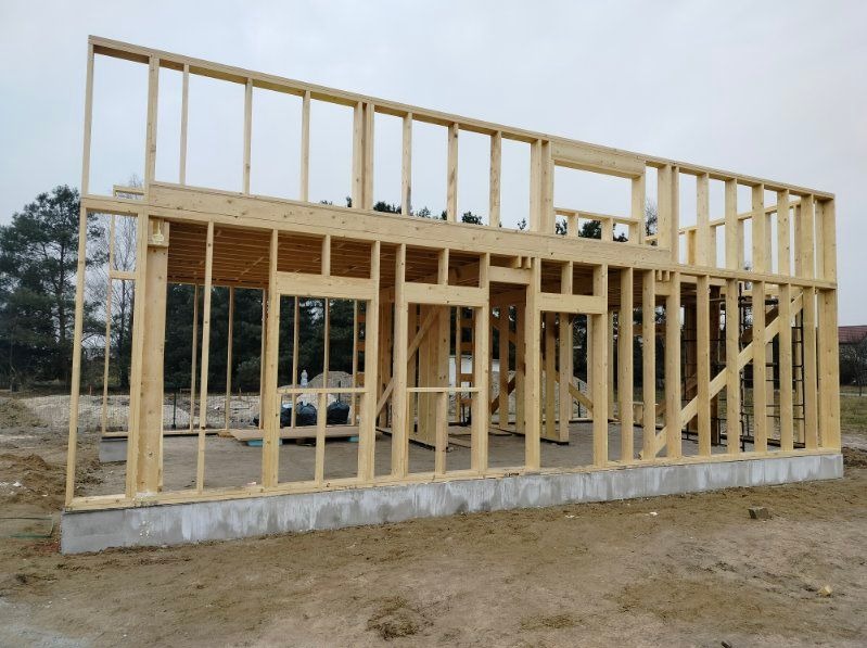 Surowa konstrukcja szkieletowa domu z drewna na betonowej podmurówce, widok z zewnątrz na tle zieleni. Elementy konstrukcyjne z jasnego drewna, widoczne okna i drzwi.