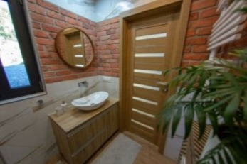 Wnętrze łazienki z ceglaną ścianą, okrągłym lustrem nad umywalką i drewnianymi drzwiami z przeszkleniami. Roślina w rogu dodaje naturalnego akcentu.