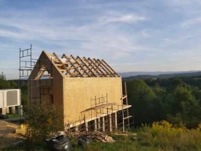 Dom w trakcie budowy z widoczną konstrukcją dachu i ścian z płyty OSB na tle zielonego krajobrazu i błękitnego nieba.