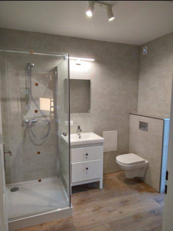 Nowoczesna łazienka z kabiną prysznicową, umywalką z szafką i podwieszaną toaletą. Szare płytki na ścianach i drewnopodobne panele podłogowe. Oświetlenie sufitowe.