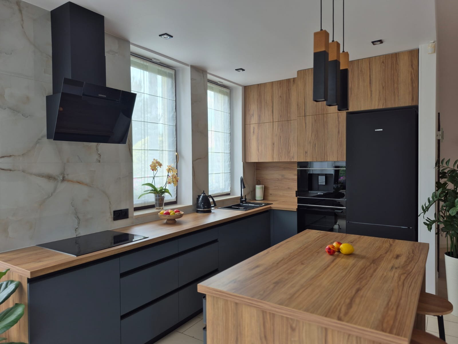 Nowoczesna kuchnia z drewnianymi blatami i czarnymi frontami szafek. Na blacie owoce, a nad nim okno z roletami rzymskimi. Czarny okap nad płytą indukcyjną.