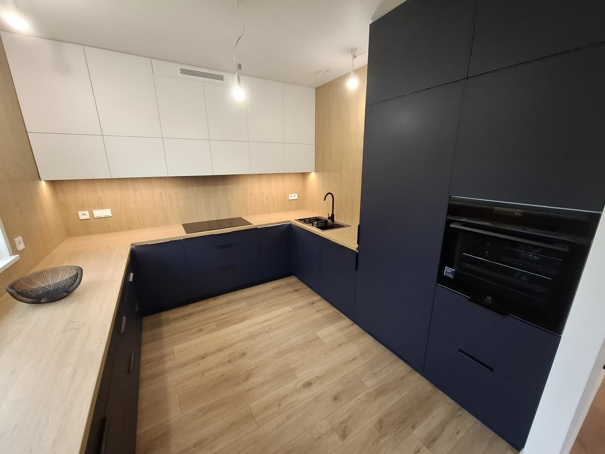 Nowoczesna kuchnia z ciemnoniebieskimi szafkami dolnymi, drewnianym blatem i białymi szafkami górnymi. Widoczne oświetlenie LED podszafkowe i designerska misa na blacie.