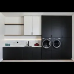 Nowoczesna pralnia: Czarne szafki z pralkami, marmurowy blat z podświetleniem LED, białe szafki wiszące i drewniane półki. Minimalistyczny design, jasne wnętrze.
