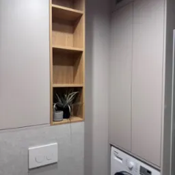 Nowoczesna łazienka: zabudowa z szafek w odcieniach szarości, drewniane półki z roślinami, pralka w zabudowie. Minimalistyczny design i funkcjonalność.