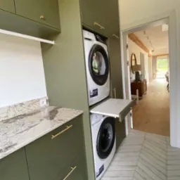 Nowoczesna pralnia w zabudowie: oliwkowe szafki z marmurowym blatem, pralka i suszarka w kolumnie z wysuwanym blatem. Podłoga w jodełkę.