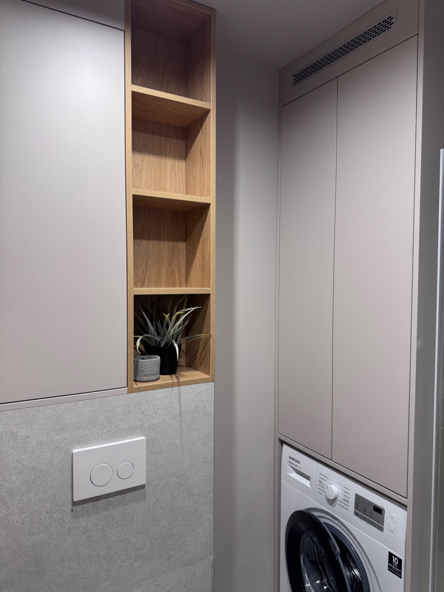 Nowoczesna łazienka: zabudowa z szafek w odcieniach szarości, drewniane półki z roślinami, pralka w zabudowie. Minimalistyczny design i funkcjonalność.