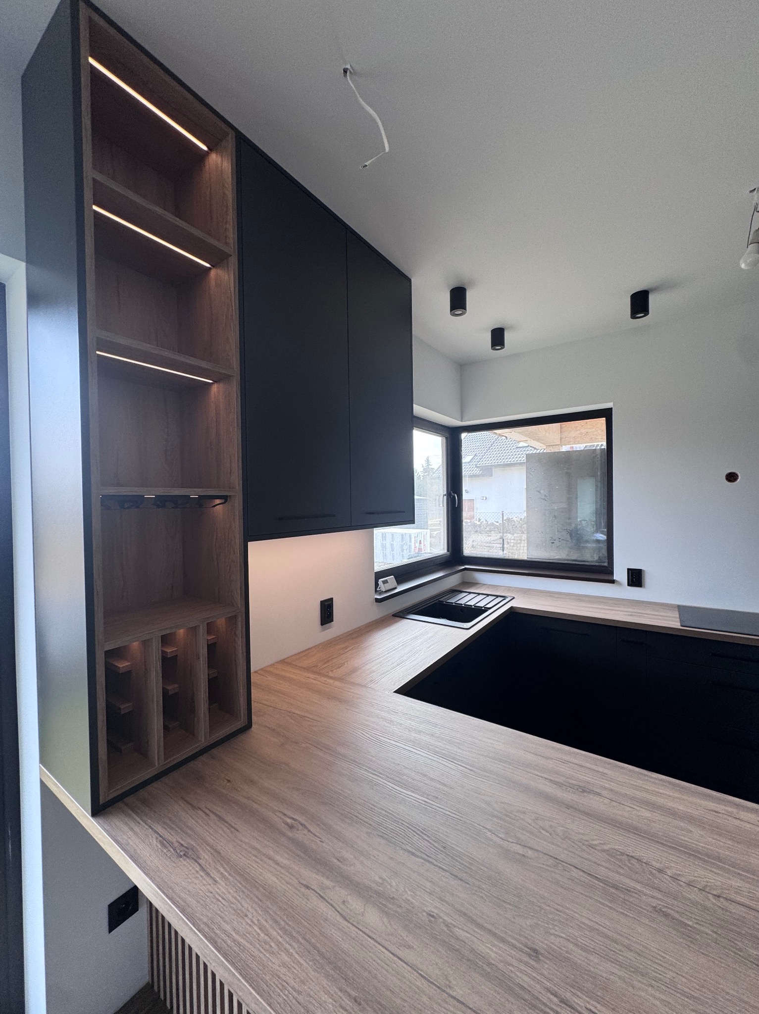 Nowoczesna kuchnia z antracytowymi szafkami i drewnopodobnym blatem. Otwarty regał z podświetleniem LED i miejscem na wino. Minimalistyczne oświetlenie sufitowe.