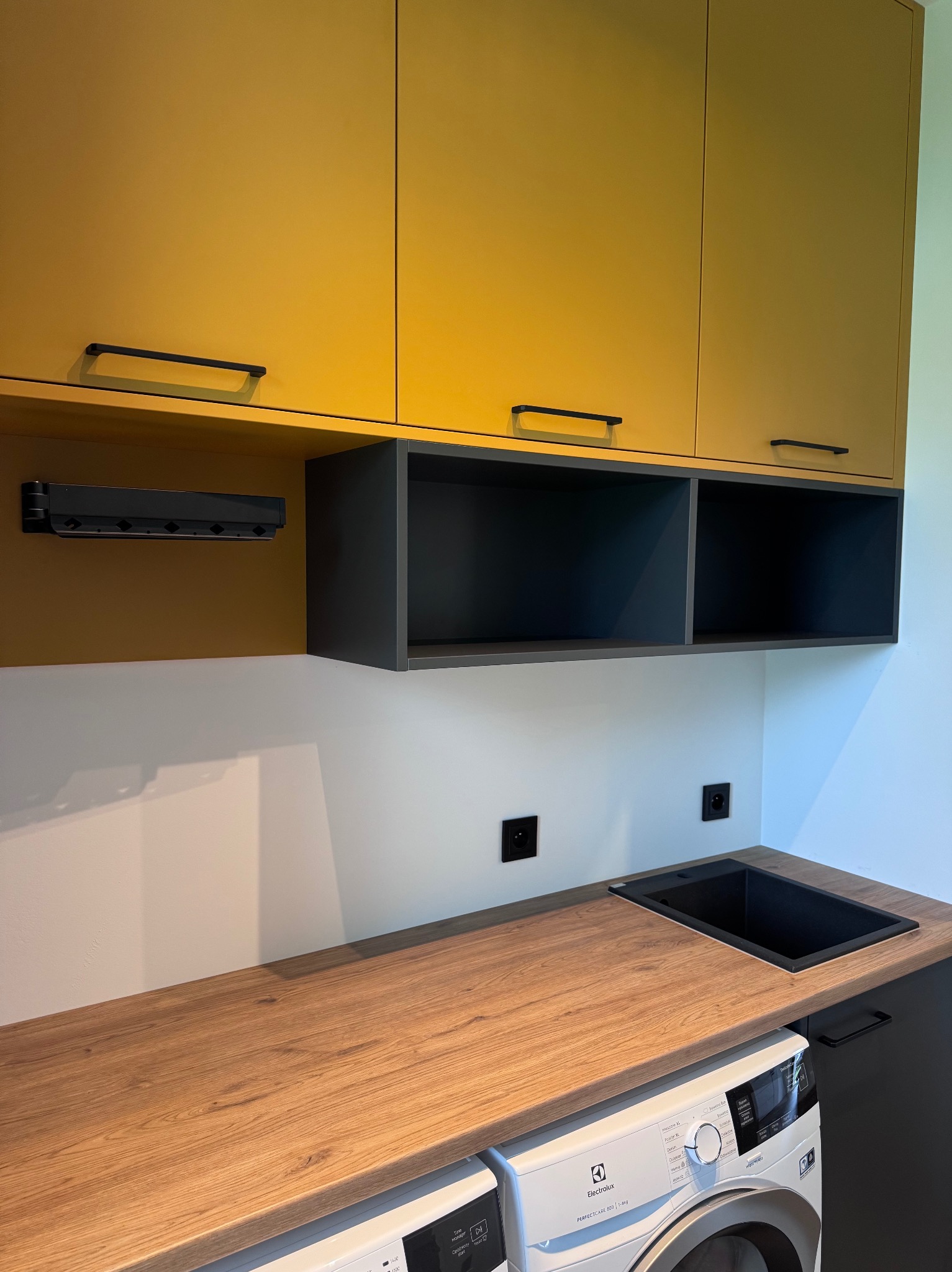 Nowoczesna aranżacja pralni: żółte szafki górne z czarnymi uchwytami, drewniany blat z czarnym zlewem i pralka. Minimalistyczny design z akcentem kolorystycznym.