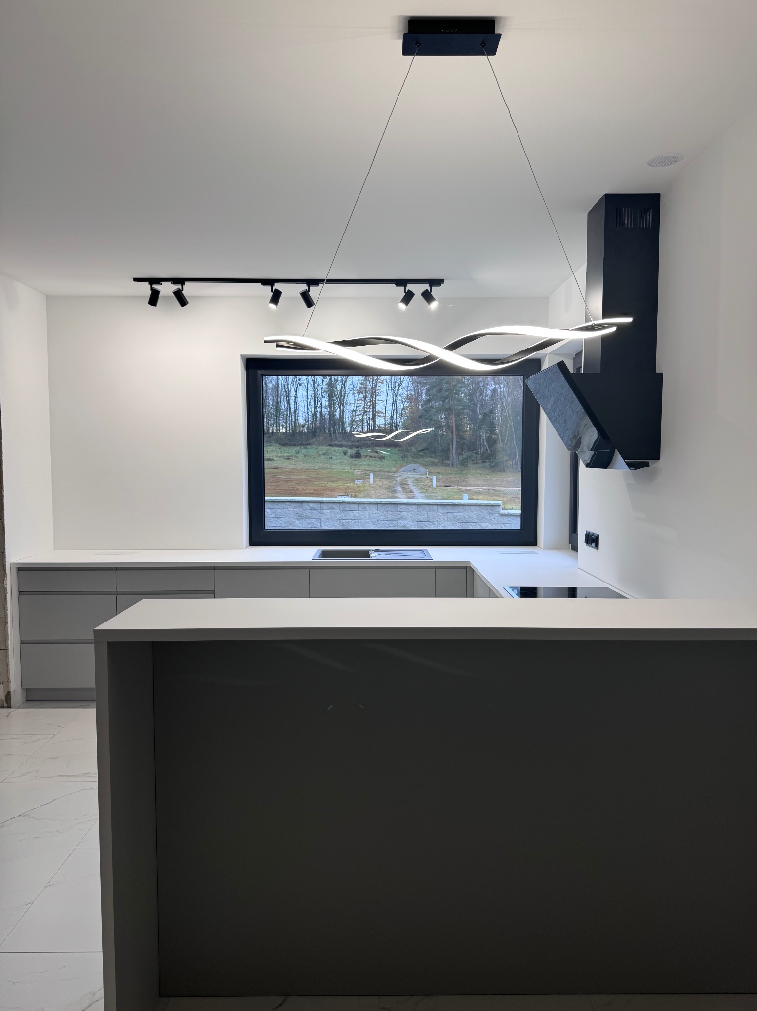 Minimalistyczna, nowoczesna kuchnia z szarymi szafkami i blatem, czarnym okapem i oknem z widokiem na zewnątrz. Oświetlenie punktowe i designerska lampa wisząca.