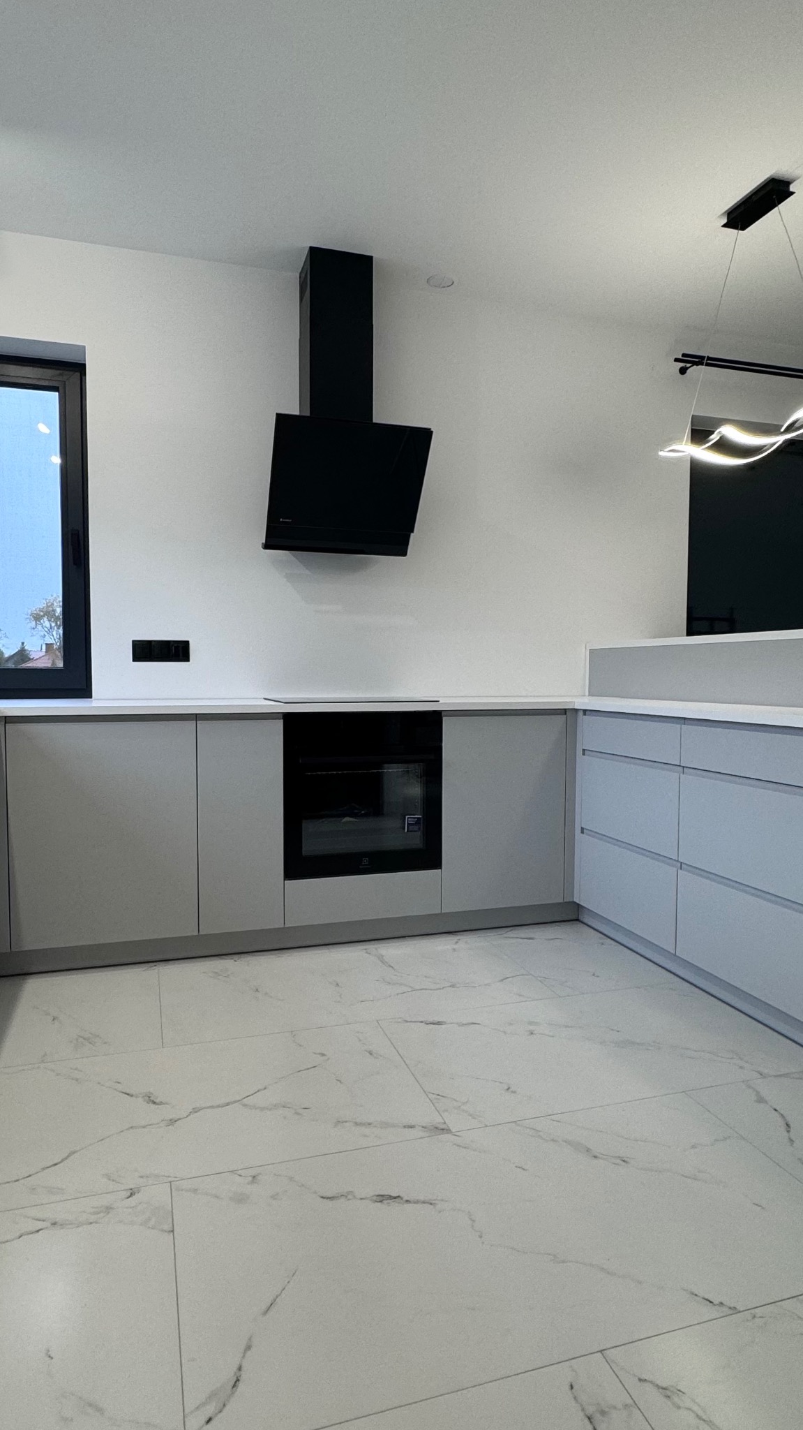 Nowoczesna kuchnia z szarymi szafkami, czarnym okapem i piekarnikiem. Biała podłoga imitująca marmur. Minimalistyczny design, oświetlenie LED.