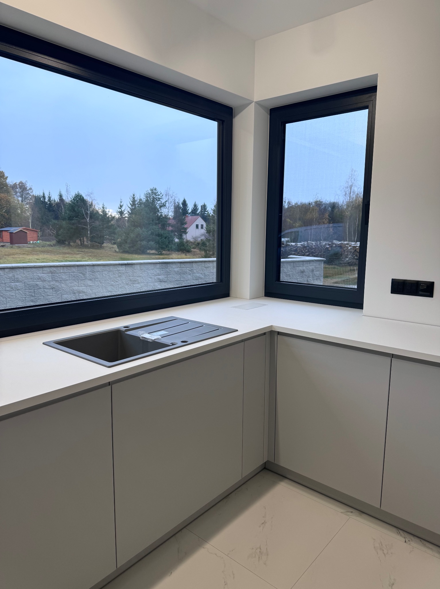 Minimalistyczna kuchnia z szarymi szafkami i blatem, zlew wpuszczony w blat, duże okna z widokiem na zieleń. Nowoczesne wykończenie, jasne płytki podłogowe.