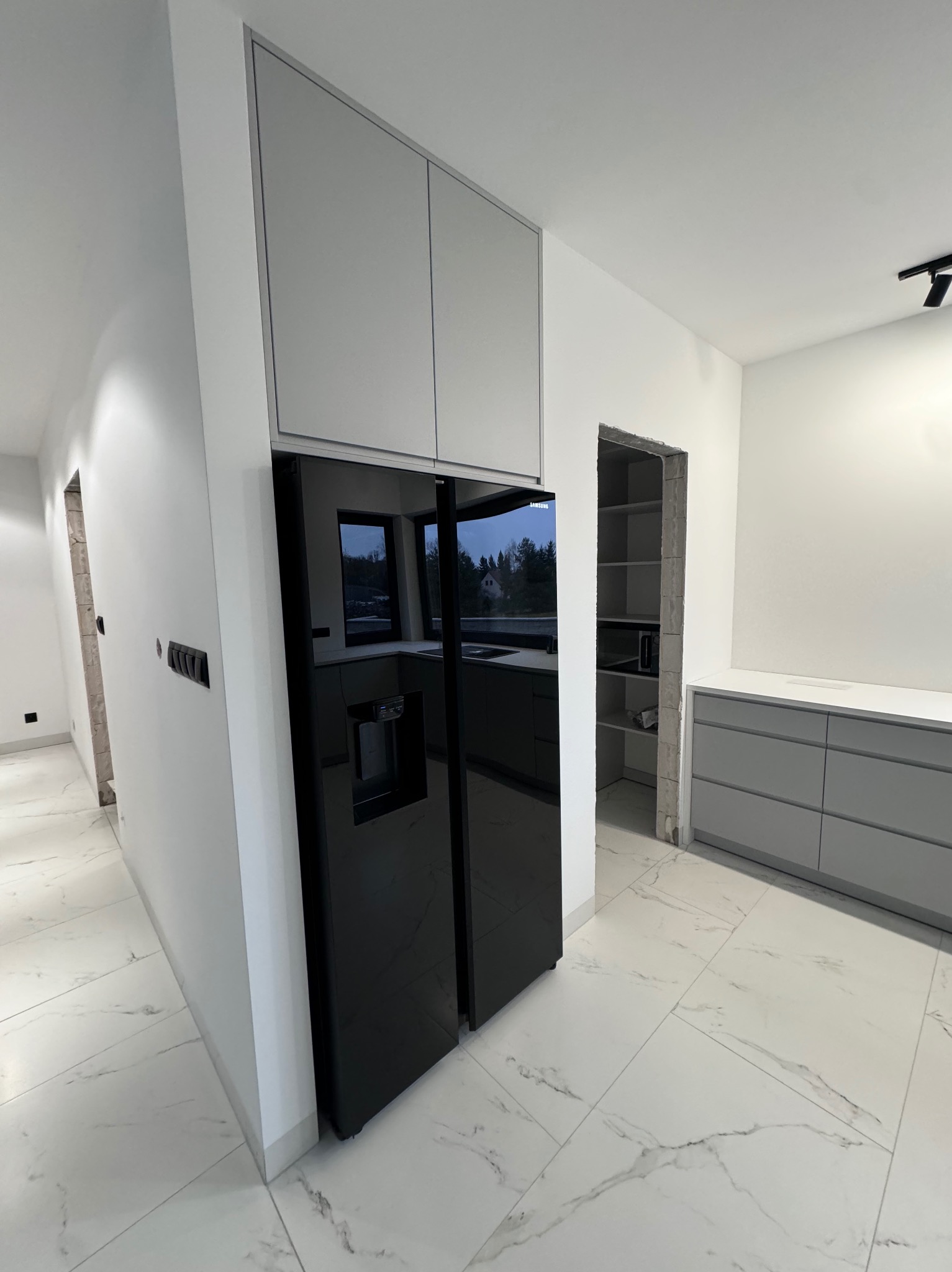 Nowoczesna kuchnia z czarną lodówką side-by-side i szarymi szafkami, biała marmurowa podłoga, widok na spiżarnię. Minimalistyczny design, oświetlenie punktowe.