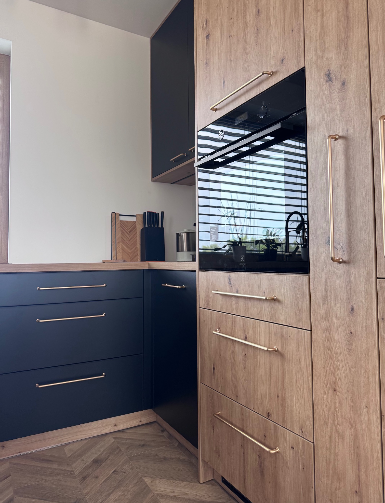 Nowoczesna kuchnia z drewnianymi i grafitowymi frontami, złotymi uchwytami i parkietem ułożonym w jodełkę. Wbudowany piekarnik odbija światło z okna.