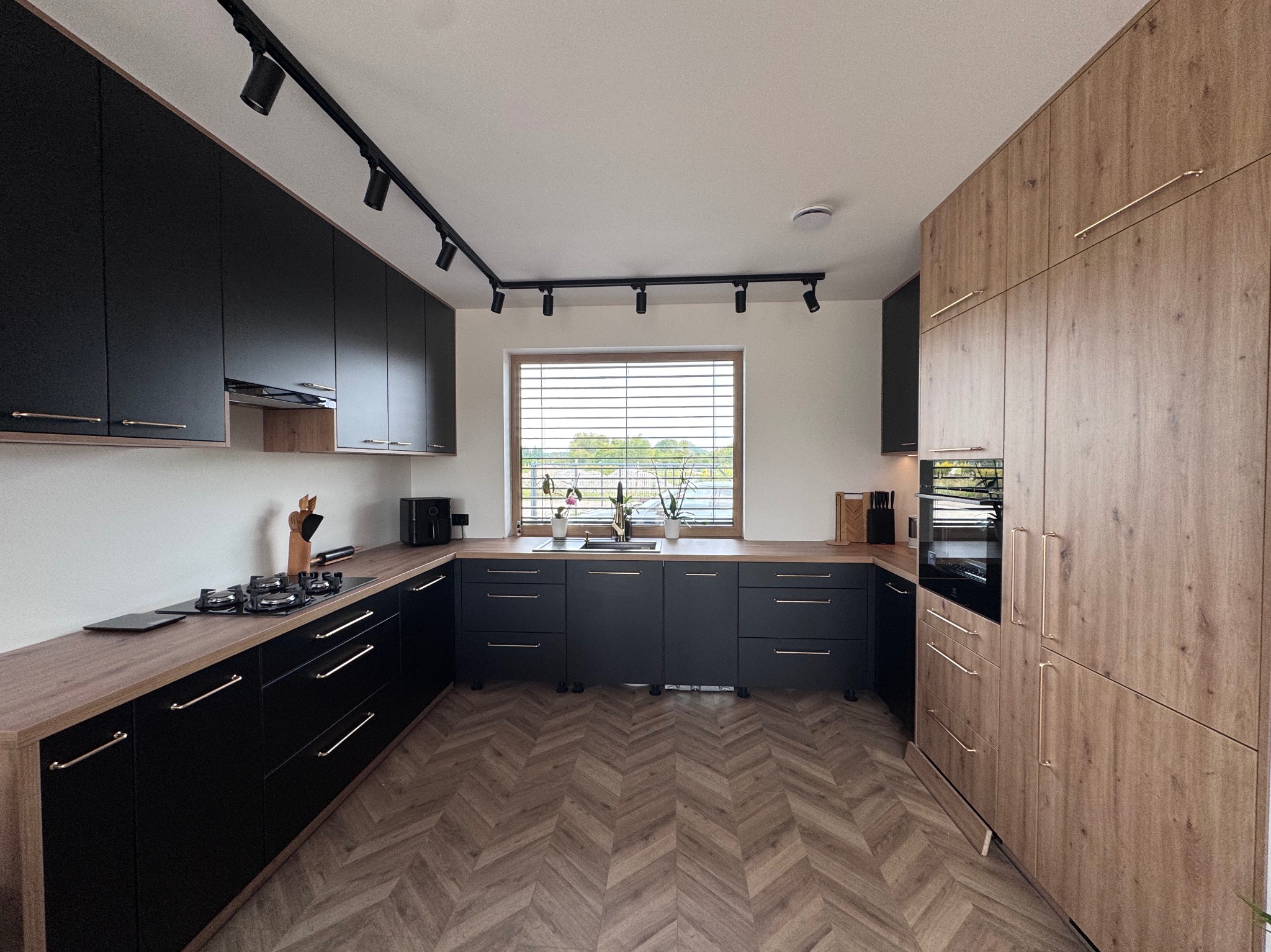 Nowoczesna kuchnia z czarnymi szafkami i drewnianym blatem, oświetlona reflektorami. Parkiet ułożony w jodełkę. Widok na okno z żaluzjami i zielenią na zewnątrz.