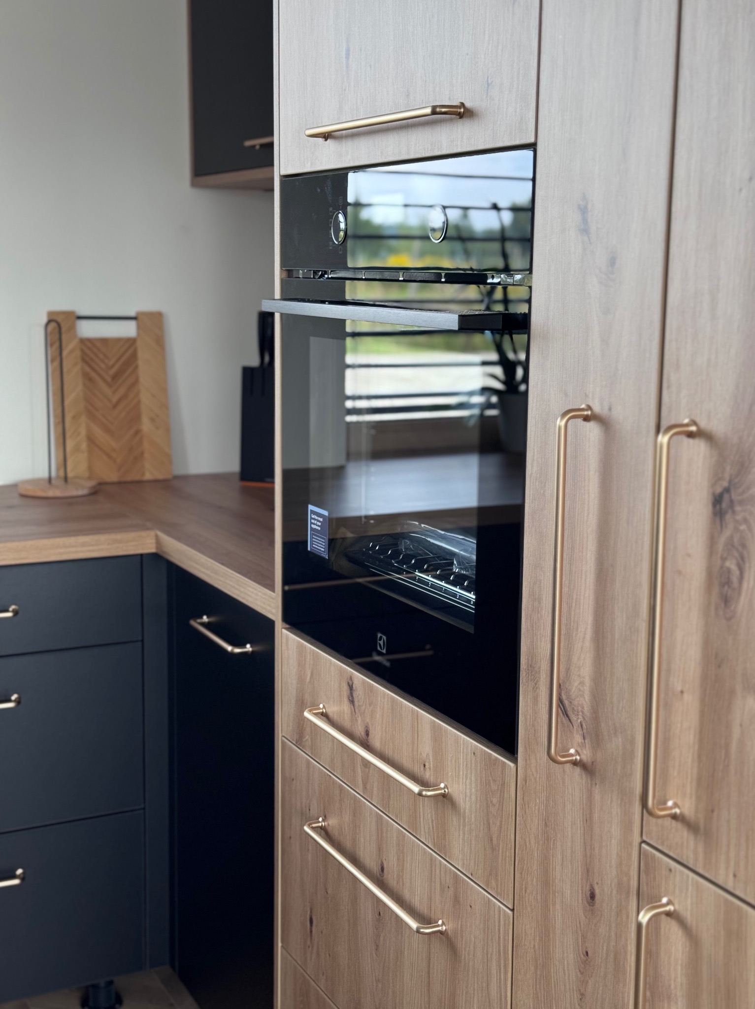 Nowoczesna zabudowa kuchenna z piekarnikiem Electrolux, fronty w odcieniach drewna i granatu, złote uchwyty. Widoczne blaty i akcesoria kuchenne.