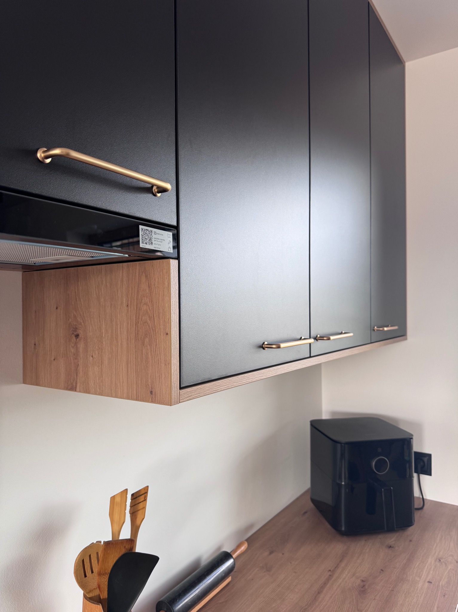 Czarne szafki kuchenne z drewnianymi akcentami i złotymi uchwytami, uzupełnione o sprzęt AGD na blacie. Minimalistyczny design.