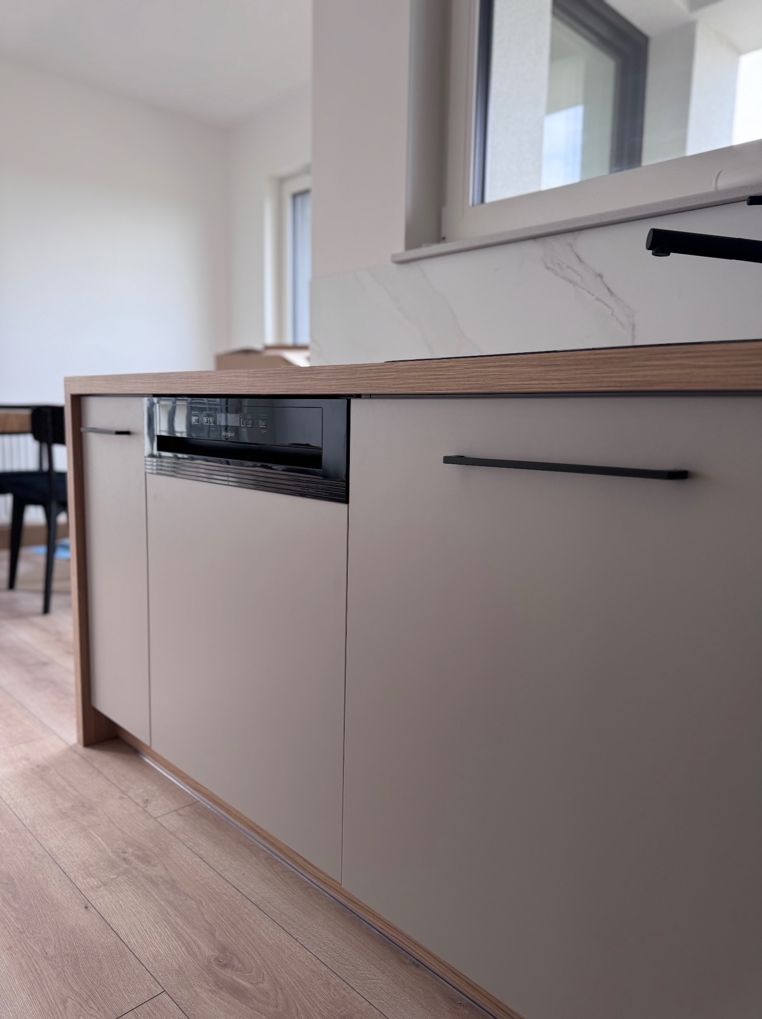 Frontalny widok na fragment zabudowy kuchennej z szarymi frontami, drewnopodobnym blatem i czarnymi uchwytami. Widoczna zintegrowana z zabudową zmywarka.