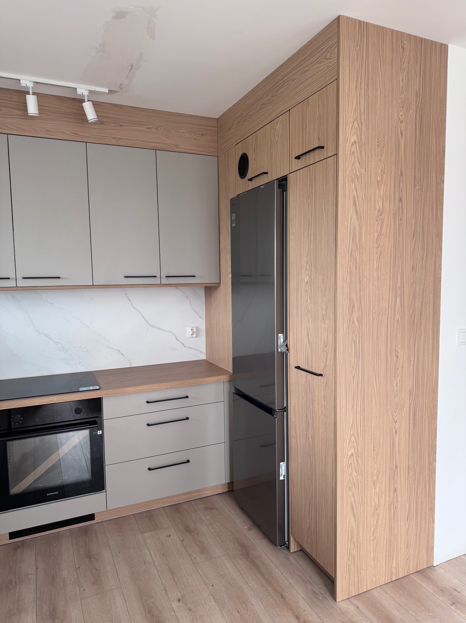 Nowoczesna kuchnia z szarymi frontami i drewnopodobnymi elementami. Widoczny piekarnik, lodówka i marmurowy backsplash. Na suficie lampy punktowe.